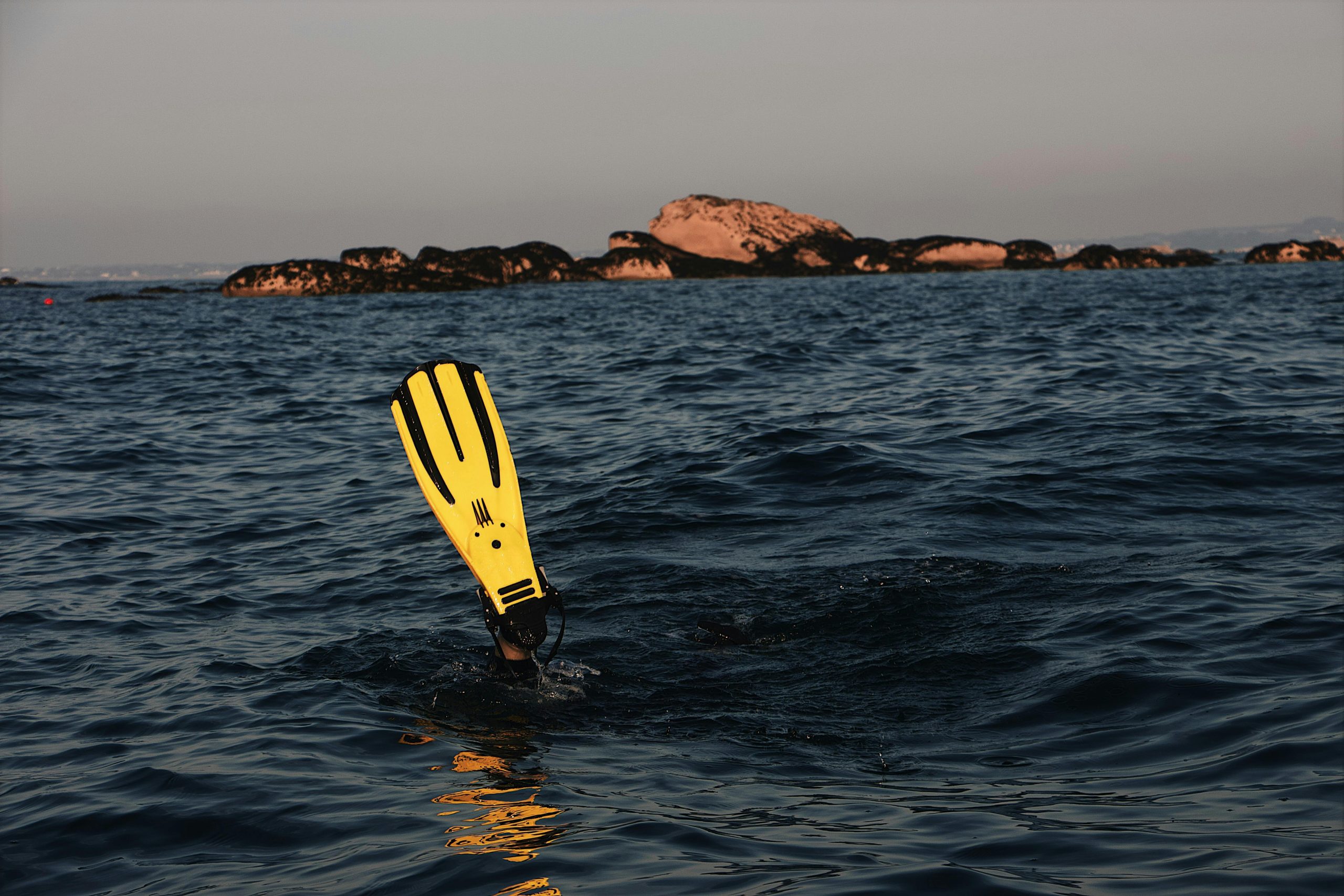 A diver in ocean waters with prominent yellow flippers, exploring near rocky islands.